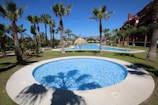 A tranquil outdoor setting featuring a circular swimming pool surrounded by palm trees and manicured grass. In the background, there is a larger pool with a thatched sunshade, creating a serene holiday atmosphere under a clear blue sky.