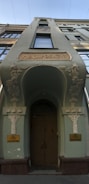 An ornate, tall building facade with decorative architectural elements featuring floral motifs. It has a central arched door with intricate carvings around it and Russian text above the entrance. The upper floors display a series of windows, and the overall structure is painted in a muted green shade.