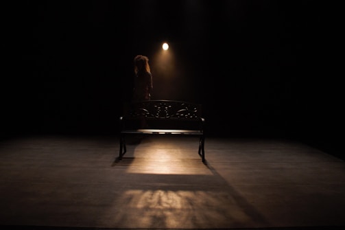 A dimly lit stage with a single spotlight illuminating a solitary metal bench. A person with curly hair stands behind the bench, partially obscured by shadow, creating a dramatic and contemplative atmosphere.
