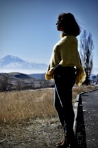 A person wearing a yellow shawl stands on a roadside, gazing towards a snow-capped mountain under a clear blue sky. The landscape features dry grass and scattered trees, with a tall, leafless tree in the background.