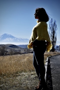 A person wearing a yellow shawl stands on a roadside, gazing towards a snow-capped mountain under a clear blue sky. The landscape features dry grass and scattered trees, with a tall, leafless tree in the background.