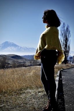 A person wearing a yellow shawl stands on a roadside, gazing towards a snow-capped mountain under a clear blue sky. The landscape features dry grass and scattered trees, with a tall, leafless tree in the background.