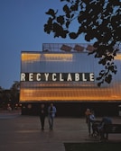 A large structure with illuminated lettering spelling 'RECYCLABLE' dominates the scene. The building's exterior is lit with warm yellow and orange lights contrasting against a dark evening sky. In the foreground, there are a few people walking and sitting near the structure, with trees silhouetted against the sky.