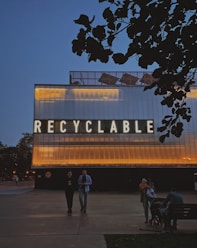 A large structure with illuminated lettering spelling 'RECYCLABLE' dominates the scene. The building's exterior is lit with warm yellow and orange lights contrasting against a dark evening sky. In the foreground, there are a few people walking and sitting near the structure, with trees silhouetted against the sky.