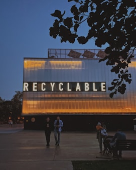 A large structure with illuminated lettering spelling 'RECYCLABLE' dominates the scene. The building's exterior is lit with warm yellow and orange lights contrasting against a dark evening sky. In the foreground, there are a few people walking and sitting near the structure, with trees silhouetted against the sky.