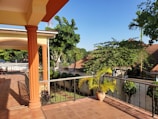 A sunny outdoor terrace with tiled flooring, featuring ornate iron railings and large, orange columns. The area is surrounded by lush green trees and plants, with a potted plant placed near the railings. In the background, rooftops of nearby buildings can be seen along with a bright blue sky.