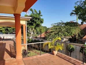 A sunny outdoor terrace with tiled flooring, featuring ornate iron railings and large, orange columns. The area is surrounded by lush green trees and plants, with a potted plant placed near the railings. In the background, rooftops of nearby buildings can be seen along with a bright blue sky.