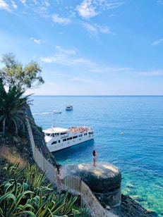 A scenic coastal view with a large white ferry boat crowded with people navigating through calm blue waters. The foreground features a rocky cliff with lush green vegetation and a girl standing on a concrete structure at the edge, taking in the view. Another small boat is visible in the background.