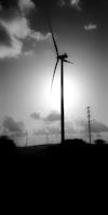 A large wind turbine stands silhouetted against the bright sun, surrounded by smaller turbines in the background. Power lines stretch across the scene, and fluffy clouds scatter across the sky.