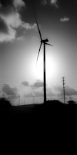 A large wind turbine stands silhouetted against the bright sun, surrounded by smaller turbines in the background. Power lines stretch across the scene, and fluffy clouds scatter across the sky.
