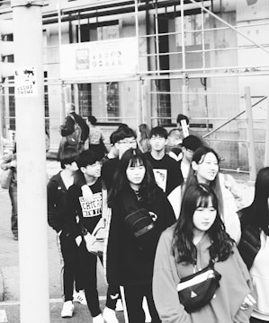 A group of young people walking together near a construction site. They appear to be casually dressed, with some wearing hoodies and jackets. The background includes scaffolding and signs, indicating construction work.