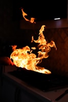 Vibrant flames rise from a sizzling pan on a stovetop, casting a warm glow in a dimly lit kitchen environment. The fire twists and dances energetically, suggesting high heat and intense cooking activity.