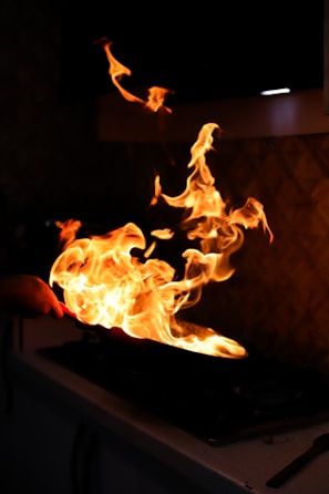 Vibrant flames rise from a sizzling pan on a stovetop, casting a warm glow in a dimly lit kitchen environment. The fire twists and dances energetically, suggesting high heat and intense cooking activity.