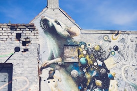 A vibrant street art mural featuring a large, realistic mouse or rat painted on the side of a building, with a whimsical, abstract background filled with planets, stars, and various intricate patterns. The artwork covers several walls of a brick structure under a clear blue sky.