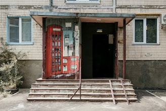 An entrance to a building featuring an artistic door painted with a London-themed design, including a red telephone box and abstract buildings. The structure includes a small set of stairs leading to the door, surrounded by tiled walls, with two windows on either side and an air conditioning unit visible on the right. Some vegetation is present on the left side of the stairs.