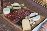 A charcuterie board featuring a variety of meats, cheeses, and accompaniments. Sliced meats are neatly arranged alongside assorted cheeses including soft white rind cheese, a slice of semi-hard cheese, and a small round of spreadable cheese. Fresh fruit such as apple slices, strawberries, and blueberries is present, along with pistachios and pecans. The background includes a textured surface and a plate of crackers.