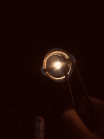 A hand holds a pair of tweezers gripping a metallic object that is glowing brightly at the center, surrounded by darkness.