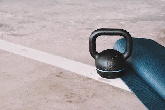 A black kettlebell rests next to a rolled-up blue yoga mat on a textured concrete floor with a white line in the background.