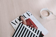 A neatly arranged still life scene with a Vogue magazine featuring a model in a red outfit, a striped notebook with a pen, and a cup of frothy coffee placed on a light wooden surface.