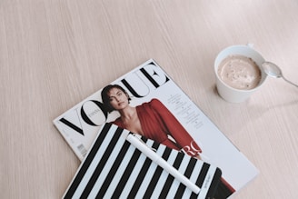 A neatly arranged still life scene with a Vogue magazine featuring a model in a red outfit, a striped notebook with a pen, and a cup of frothy coffee placed on a light wooden surface.