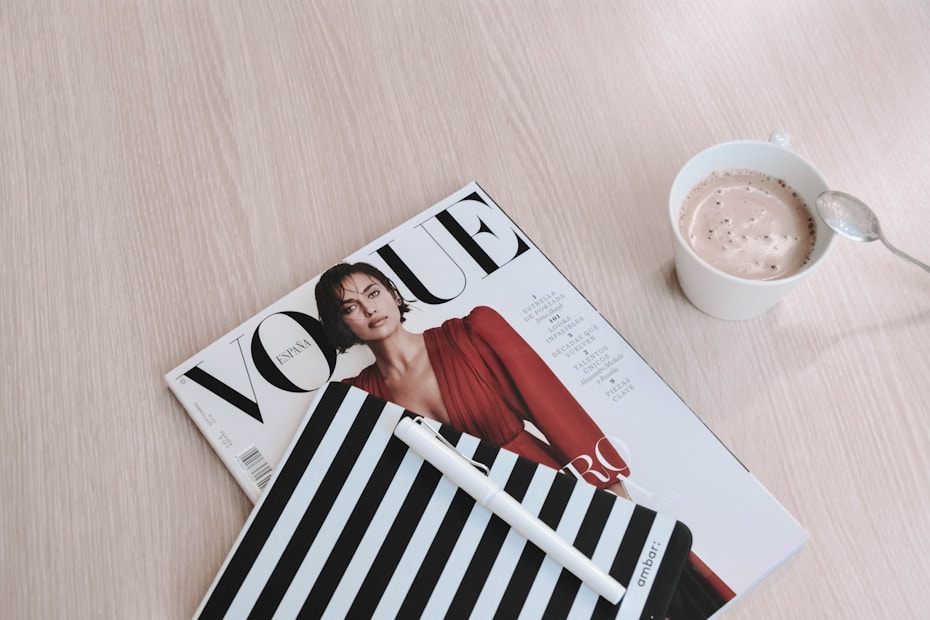 A neatly arranged still life scene with a Vogue magazine featuring a model in a red outfit, a striped notebook with a pen, and a cup of frothy coffee placed on a light wooden surface.