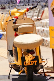 A yellow vintage scooter with a worn seat is parked on a street, marked with the letters GRPT on the rear. The scooter's handlebars are wrapped with red straps, and there are festive elements attached to it. The surrounding area contains other scooters, creating a bustling, vintage vibe.