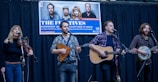 A musical group is performing on a stage with four members playing instruments, including a violin, a guitar, and a banjo. A promotional banner hangs above them with images of the band and event details.