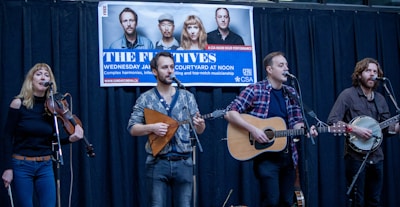 A musical group is performing on a stage with four members playing instruments, including a violin, a guitar, and a banjo. A promotional banner hangs above them with images of the band and event details.