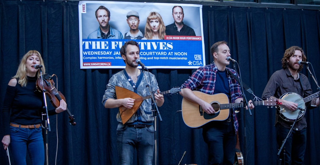 A musical group is performing on a stage with four members playing instruments, including a violin, a guitar, and a banjo. A promotional banner hangs above them with images of the band and event details.