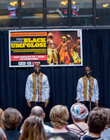 A group of performers stand on stage, each in matching traditional attire, consisting of white shirts with decorative patterns, while holding microphones. They are facing an audience seated in front of them. Behind the performers, a banner advertises a Black History Month event featuring Black Umfolosi from Zimbabwe. The setting is indoors, featuring a backdrop of dark curtains and a partially visible upper gallery.