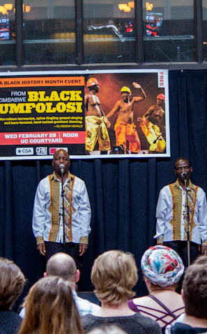A group of performers stand on stage, each in matching traditional attire, consisting of white shirts with decorative patterns, while holding microphones. They are facing an audience seated in front of them. Behind the performers, a banner advertises a Black History Month event featuring Black Umfolosi from Zimbabwe. The setting is indoors, featuring a backdrop of dark curtains and a partially visible upper gallery.