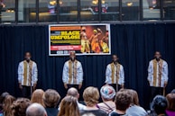 A group of performers stand on stage, each in matching traditional attire, consisting of white shirts with decorative patterns, while holding microphones. They are facing an audience seated in front of them. Behind the performers, a banner advertises a Black History Month event featuring Black Umfolosi from Zimbabwe. The setting is indoors, featuring a backdrop of dark curtains and a partially visible upper gallery.
