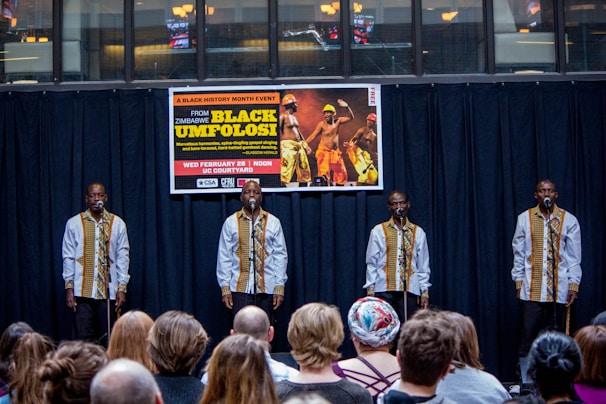 A group of performers stand on stage, each in matching traditional attire, consisting of white shirts with decorative patterns, while holding microphones. They are facing an audience seated in front of them. Behind the performers, a banner advertises a Black History Month event featuring Black Umfolosi from Zimbabwe. The setting is indoors, featuring a backdrop of dark curtains and a partially visible upper gallery.