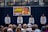 A group of performers stand on stage, each in matching traditional attire, consisting of white shirts with decorative patterns, while holding microphones. They are facing an audience seated in front of them. Behind the performers, a banner advertises a Black History Month event featuring Black Umfolosi from Zimbabwe. The setting is indoors, featuring a backdrop of dark curtains and a partially visible upper gallery.