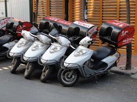 Four white scooters with black handlebars are lined up in a row, each with a red delivery box on the rear. The boxes are branded with a recognizable fast-food logo and contact information. They are parked on a paved street next to a wooden fence and a tree.