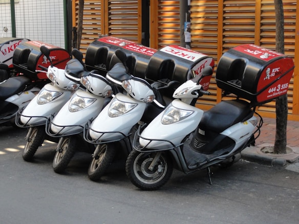 Four white scooters with black handlebars are lined up in a row, each with a red delivery box on the rear. The boxes are branded with a recognizable fast-food logo and contact information. They are parked on a paved street next to a wooden fence and a tree.