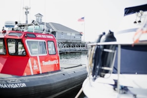 A red TowBoatU.S. rescue boat is docked next to another vessel at a marina. In the background, a building with a sign reading 'Dockside Cafe' is visible along with a flagpole displaying the American flag. The scene is bright and serene, suggesting a peaceful day by the water.