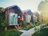Several wooden cabins with thatched roofs are lined up along a pathway, surrounded by lush greenery. Clothes are hanging outside the cabins, suggesting a laid-back, vacation atmosphere. Sunlight filters through the trees, casting a warm glow over the scene.