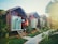 Several wooden cabins with thatched roofs are lined up along a pathway, surrounded by lush greenery. Clothes are hanging outside the cabins, suggesting a laid-back, vacation atmosphere. Sunlight filters through the trees, casting a warm glow over the scene.