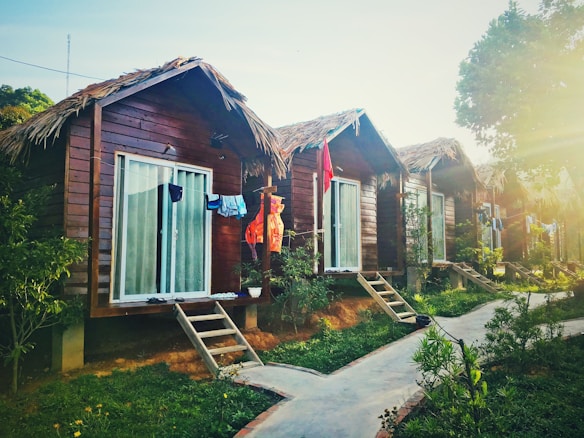 Several wooden cabins with thatched roofs are lined up along a pathway, surrounded by lush greenery. Clothes are hanging outside the cabins, suggesting a laid-back, vacation atmosphere. Sunlight filters through the trees, casting a warm glow over the scene.