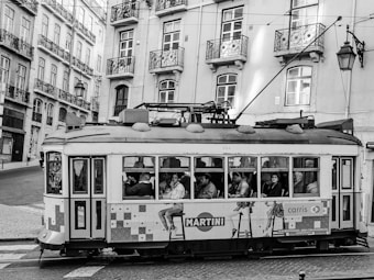 A vintage streetcar filled with passengers is traveling through a European city. The streetcar is decorated with colorful advertisements and is set against a backdrop of classical buildings with balconies and ornate windows. The scene captures a moment of daily commute with people sitting and looking out of the streetcar windows. The image is in black and white, adding a timeless and historical feel to it.