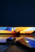 A close-up view of a modern electronic setup featuring a laptop with a colorful screen, a smartwatch, and a smartphone placed on a wooden surface. The smartphone displays a reflective surface with a prominent logo.