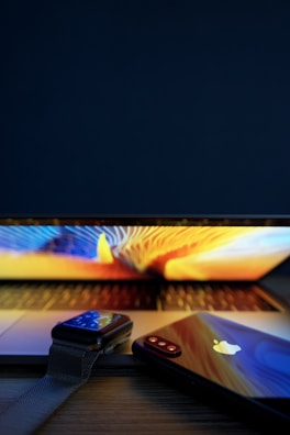 A close-up view of a modern electronic setup featuring a laptop with a colorful screen, a smartwatch, and a smartphone placed on a wooden surface. The smartphone displays a reflective surface with a prominent logo.