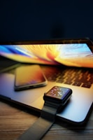 A laptop with a colorful display is open on a wooden surface, with a smartphone placed on top of it. There is also a smartwatch with a dark strap resting on the laptop's keyboard. The screen of the smartwatch displays the time with a digital watch face.