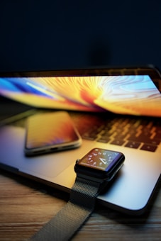 A laptop with a colorful display is open on a wooden surface, with a smartphone placed on top of it. There is also a smartwatch with a dark strap resting on the laptop's keyboard. The screen of the smartwatch displays the time with a digital watch face.