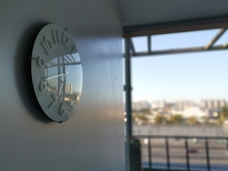 A wall-mounted clock with reflective glass showing a faint reflection of a distant cityscape. The clock has an abstract design with numbers that appear to be slightly raised. In the background, there is an out-of-focus view of a balcony overlooking a blurred urban landscape under a clear sky.