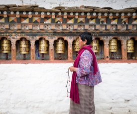A person wearing a colorful jacket and maroon scarf holds prayer beads while standing in front of a row of intricately decorated prayer wheels set into a white brick wall. The prayer wheels feature elaborate designs and inscriptions.