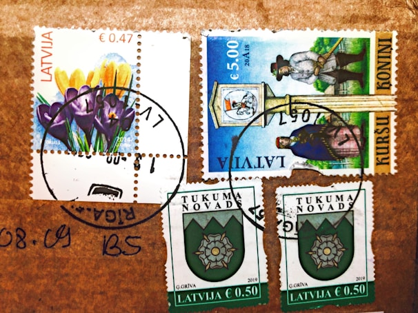 Several postage stamps from Latvia are affixed to a brown surface. The stamps feature various designs, including colorful flowers, a historical figure, and a crest emblem. Postmarks are visible, indicating the stamps have been used.