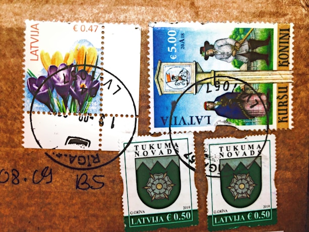 Several postage stamps from Latvia are affixed to a brown surface. The stamps feature various designs, including colorful flowers, a historical figure, and a crest emblem. Postmarks are visible, indicating the stamps have been used.