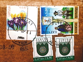 Several postage stamps from Latvia are affixed to a brown surface. The stamps feature various designs, including colorful flowers, a historical figure, and a crest emblem. Postmarks are visible, indicating the stamps have been used.